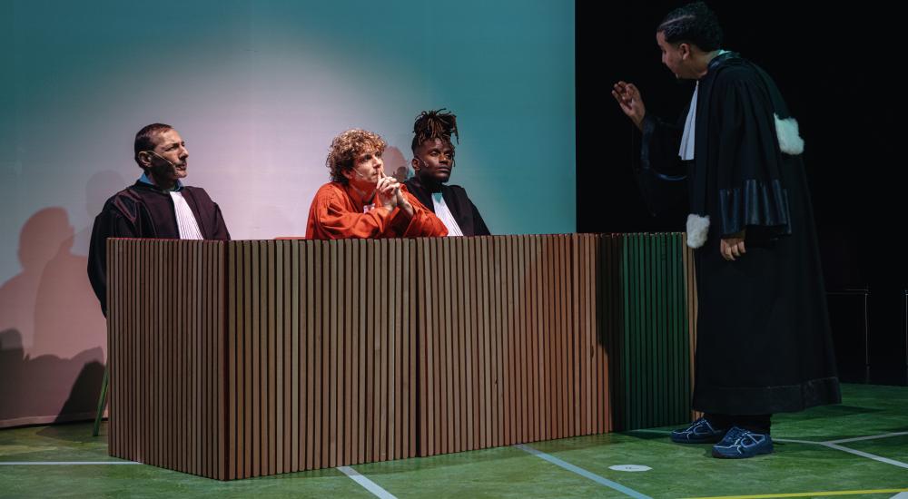 An actor dressed as a lawyer addressing three other actors, one in an orange jumpsuit and the other two also dressed as lawyers