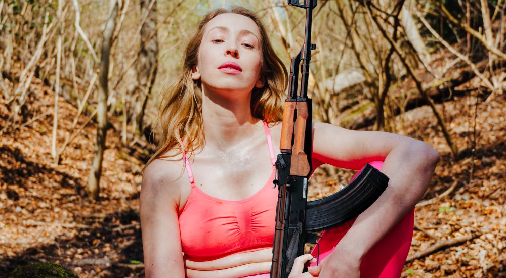 A blonde woman in yoga clothes holding an automatic rifle