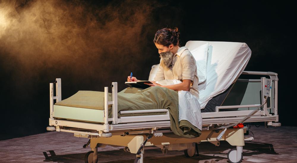 An actor wearing a fake beard writing in a hospital bed