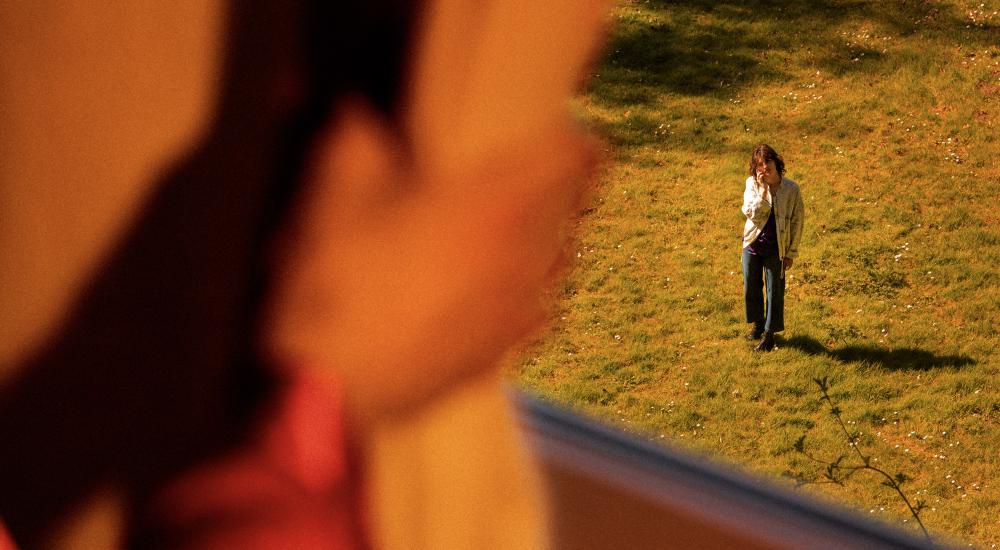 A hand holding open a curtain revealing a person standing outside on the grass