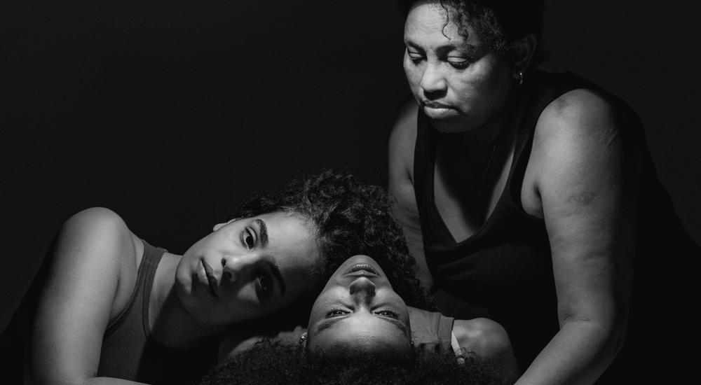 A black-and-white photo of three women holding each other, one sitting, one lying on her back with the last one resting her head on her