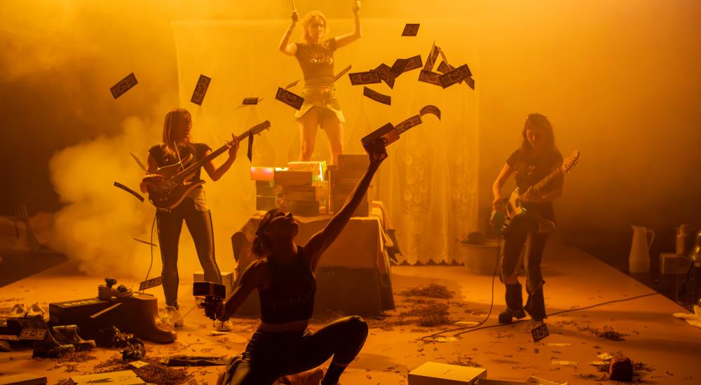 Four people on stage, two playing guitar, one holding drumsticks and the person in front shooting fake money 