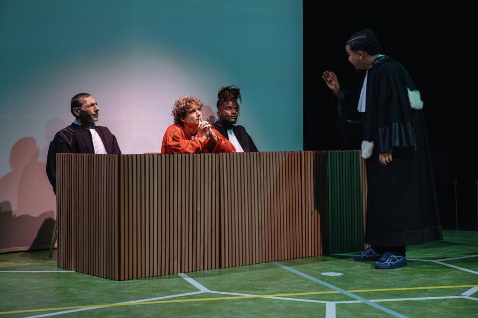 An actor dressed as a lawyer addressing three other actors, one in an orange jumpsuit and the other two also dressed as lawyers