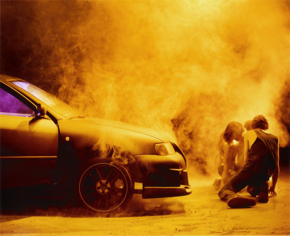Two people kneeling next to a burning car