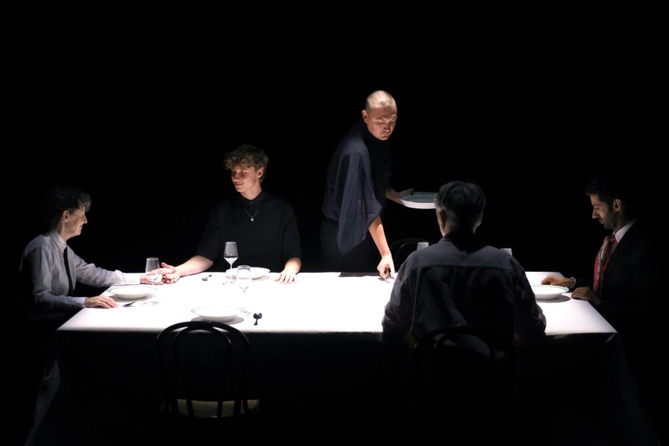 Five actors standing/sitting around a brightly lit table