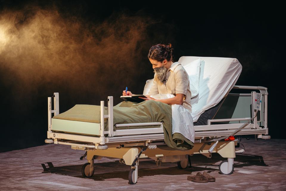 An actor wearing a fake beard writing in a hospital bed