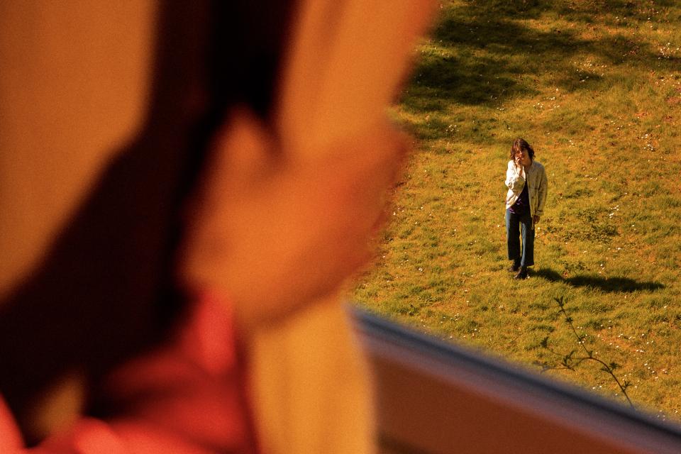 A hand holding open a curtain revealing a person standing outside on the grass