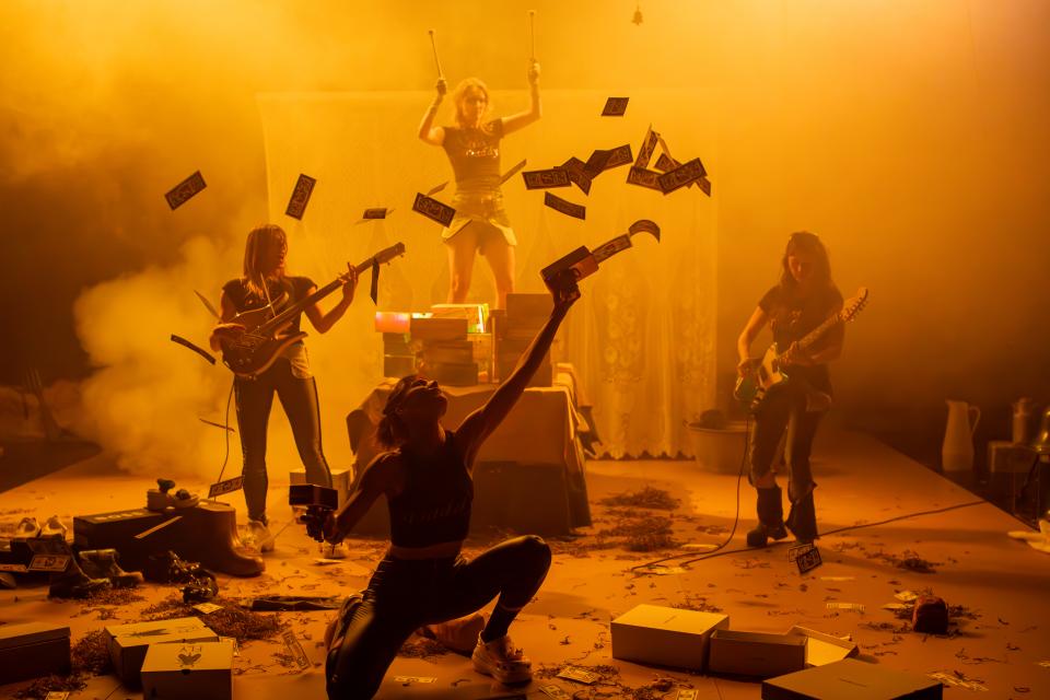 Four people on stage, two playing guitar, one holding drumsticks and the person in front shooting fake money 